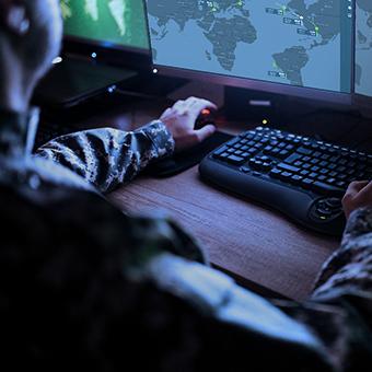 一个军人坐在台式电脑前，看着显示器上的世界地图